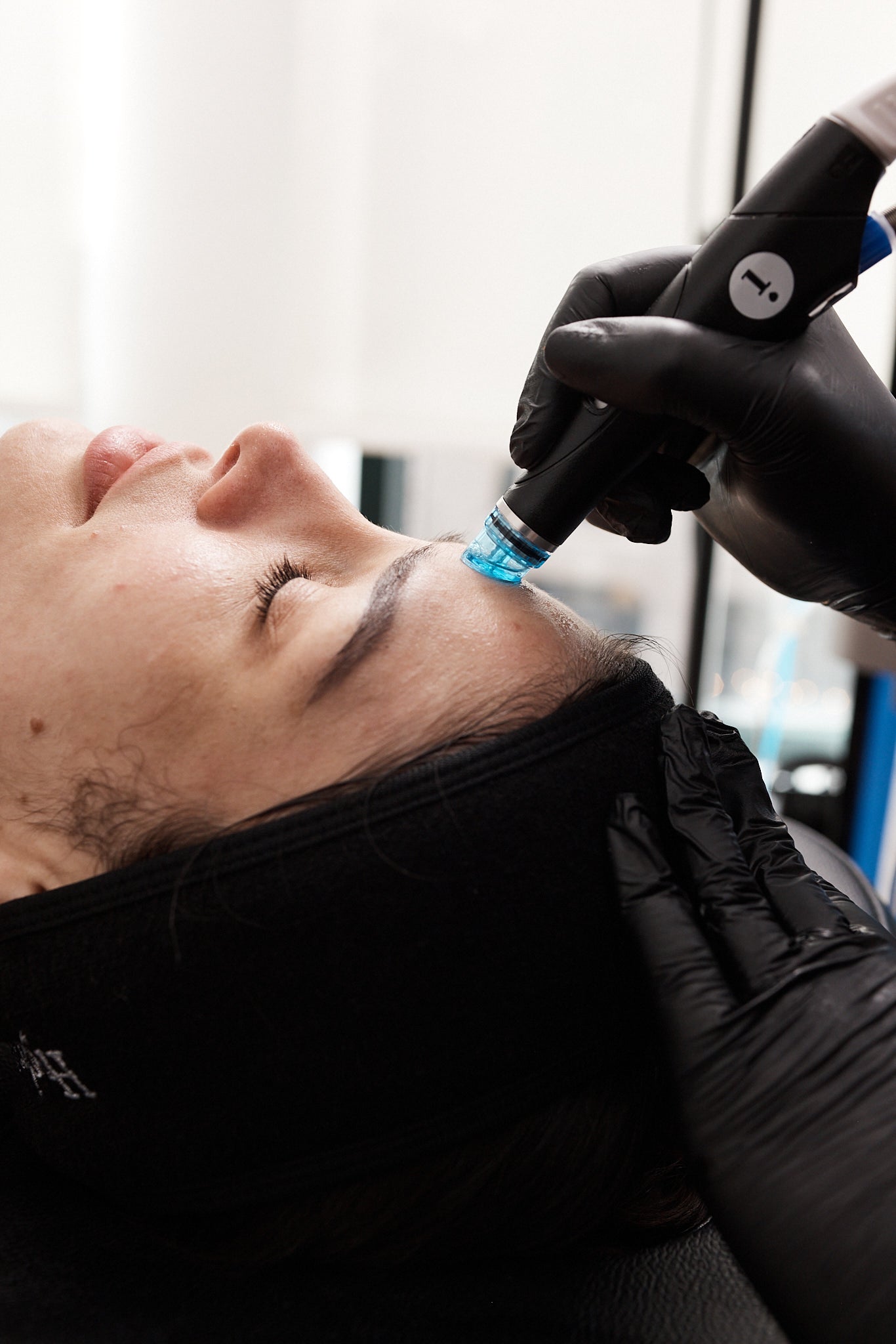 Close-up of client lying back while technician in black gloves applies Hydrafacial handpiece to forehead.
