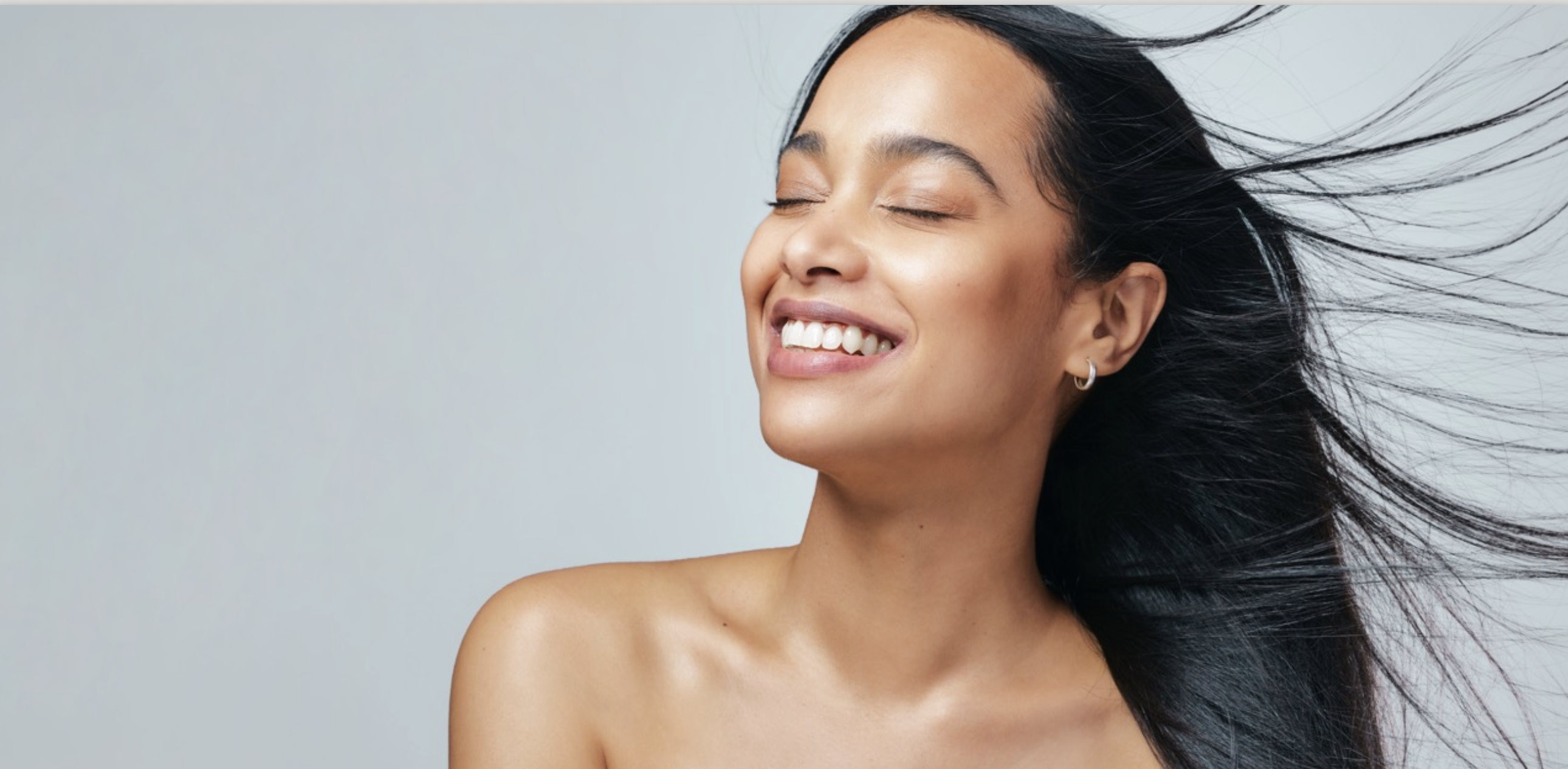 Woman smiling with eyes closed, head tilted back slightly, with long dark hair blowing behind her.