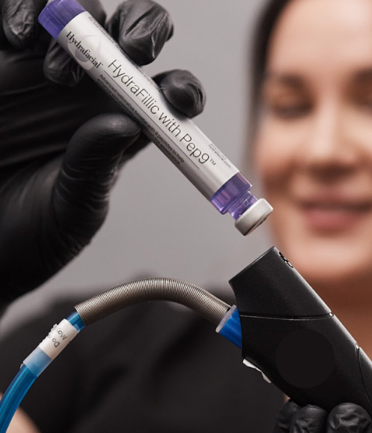Close-up of a provider holding a Hydrafacial booster vial labeled “HydraFillic with Pep9™” while preparing to load it into the handheld device.