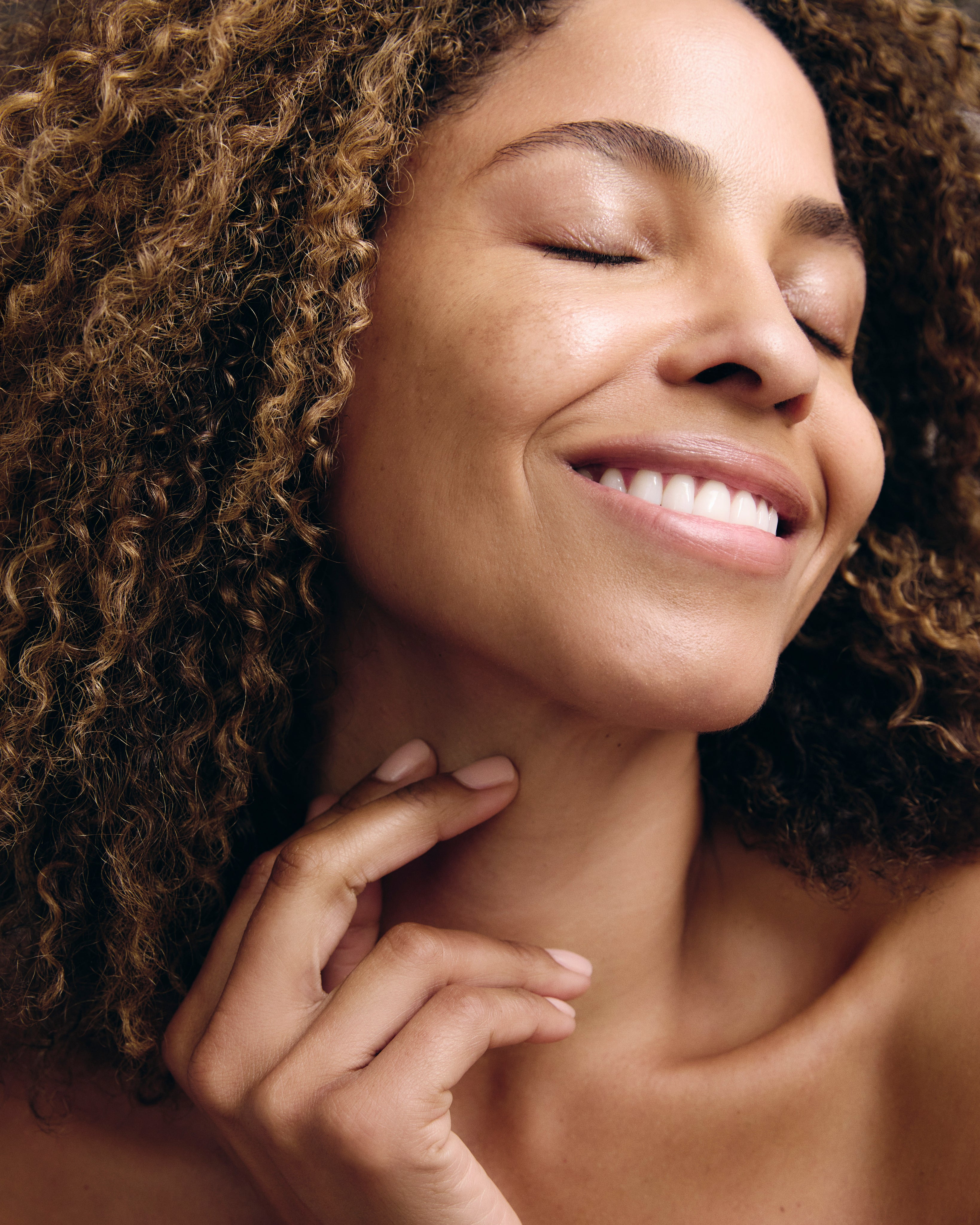 A woman with curly hair smiling with eyes closed, showing clear, luminous skin and a relaxed expression.