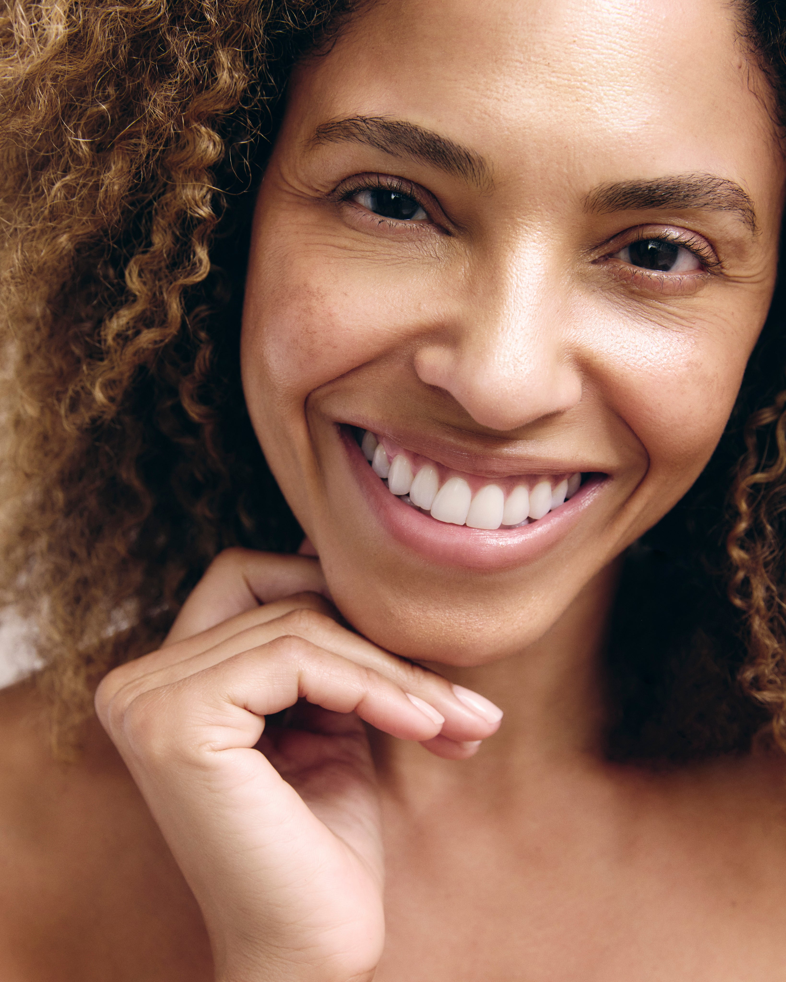 A close-up of a curly-haired woman smiling with her hand resting under her chin.