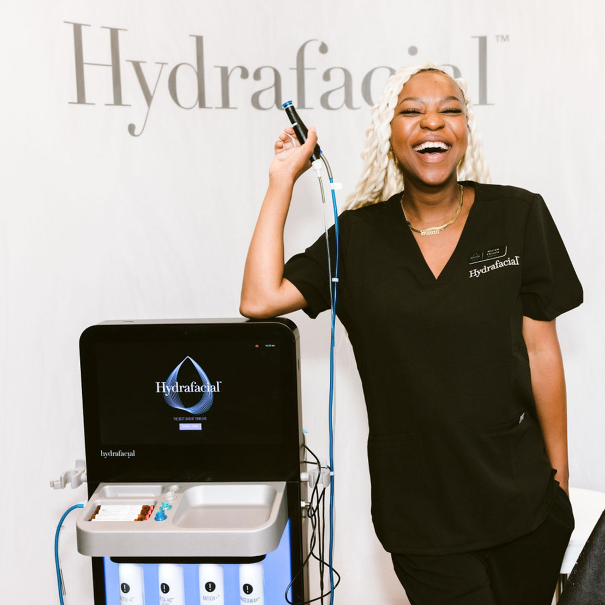 Hydrafacial provider in uniform smiling while holding the device next to a Syndeo machine.