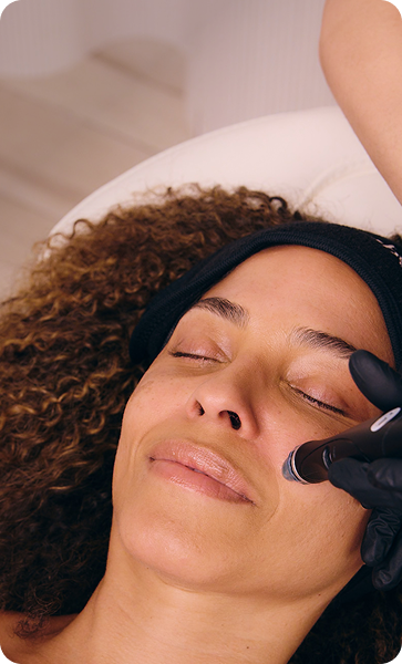 A curly-haired woman lies back with her eyes closed as a Hydrafacial device is applied to her cheek by a practitioner in black gloves.