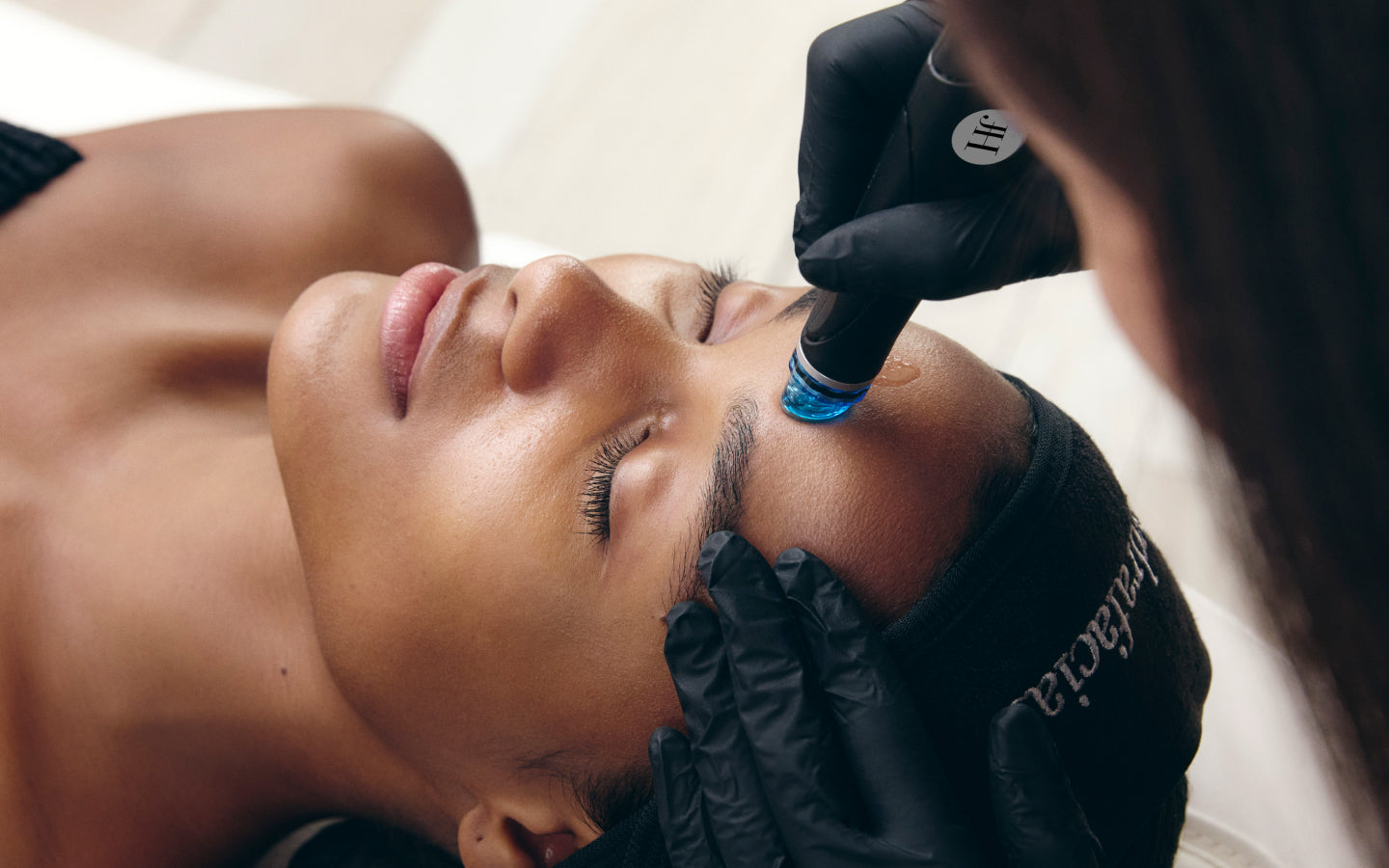 A client reclines with eyes closed while a practitioner applies a Hydrafacial device with a blue tip to the forehead area. 