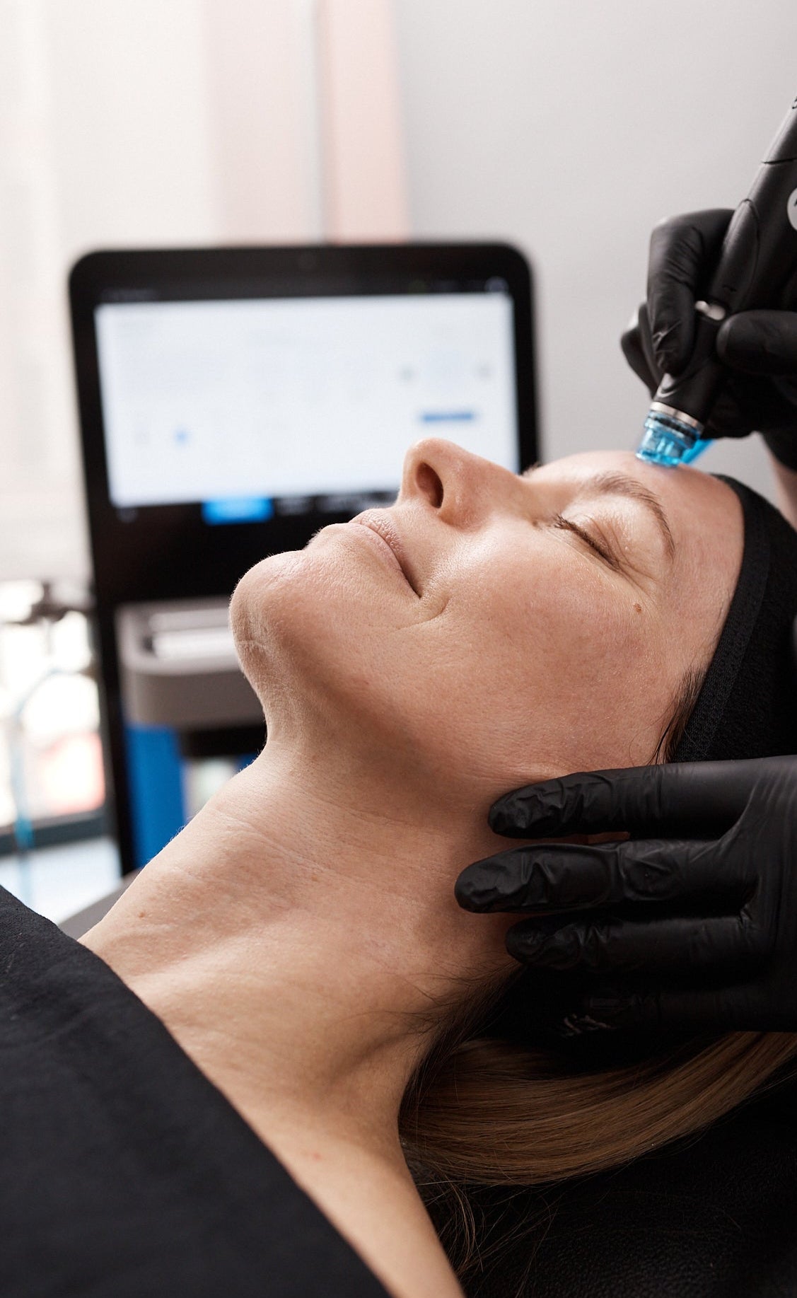 A gloved hand holds a device with a blue tip against the cheekbone near the nose of a person lying back with eyes closed.