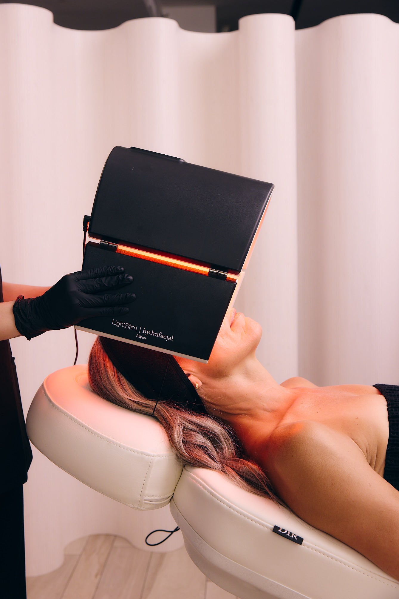 Client reclines under a Hydrafacial LightStim red LED panel while a provider adjusts the device for targeted skin rejuvenation.