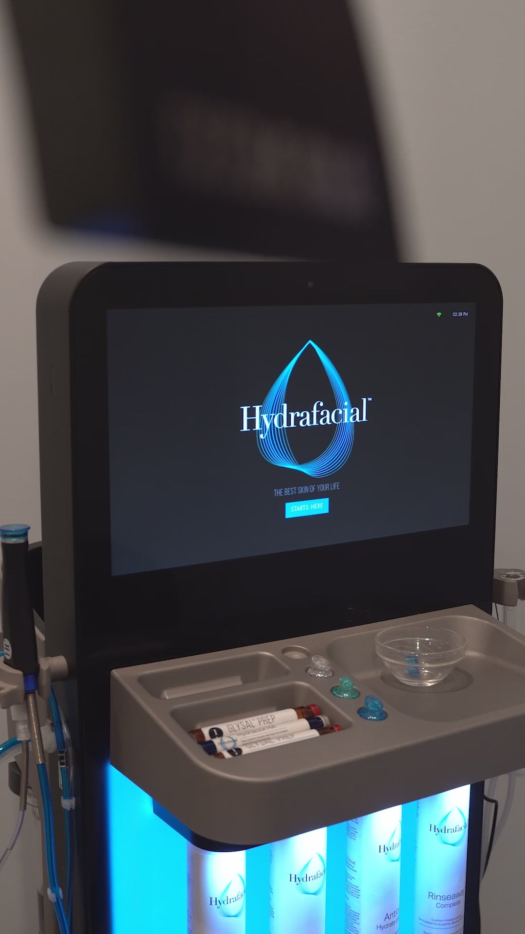 Hydrafacial treatment device displayed in a clinic setting. The screen shows the Hydrafacial logo and tagline, “The Best Skin of Your Life Starts Here.” Serums and tips are visible in the tray below.