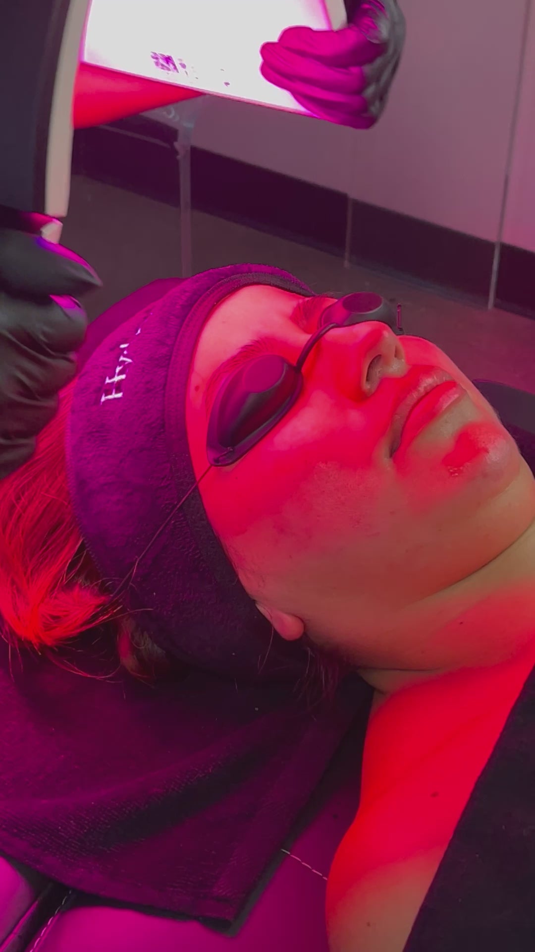 A client lies back wearing protective goggles while receiving LED light therapy during a Hydrafacial. Red light illuminates the face, targeting skin rejuvenation and calming inflammation.