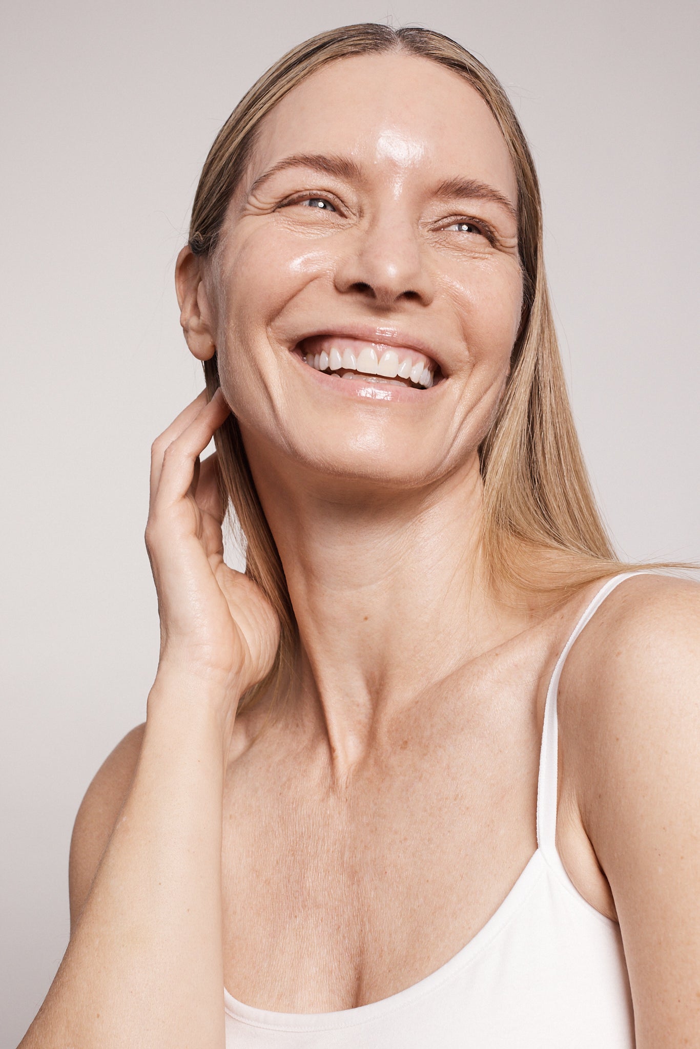 A close-up of a woman with blonde hair smiling with her hand gently touching the side of her face near the eye.